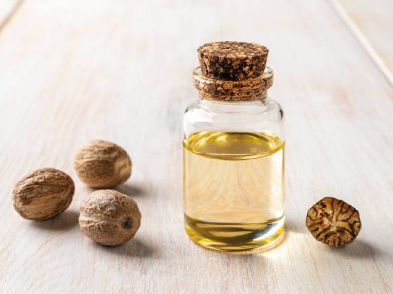 Glass jar of nutmeg essential oil closeup. Nutmeg fragrant oil and seeds in a small bottle over white wooden background. Myristica fragrans tree fruit for Ayurveda and herbal medicine. Front view.